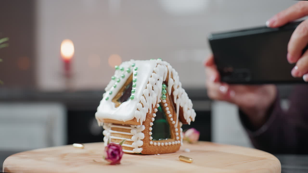스마트폰을 들고 있는 손의 클로즈업으로 나무 트레이에 장식된 진저브레드 집을 촬영합니다. 배경에는 따뜻한 불빛, 부드러운 흐릿함, 그리고 아한 부 분위기가 있습니다.