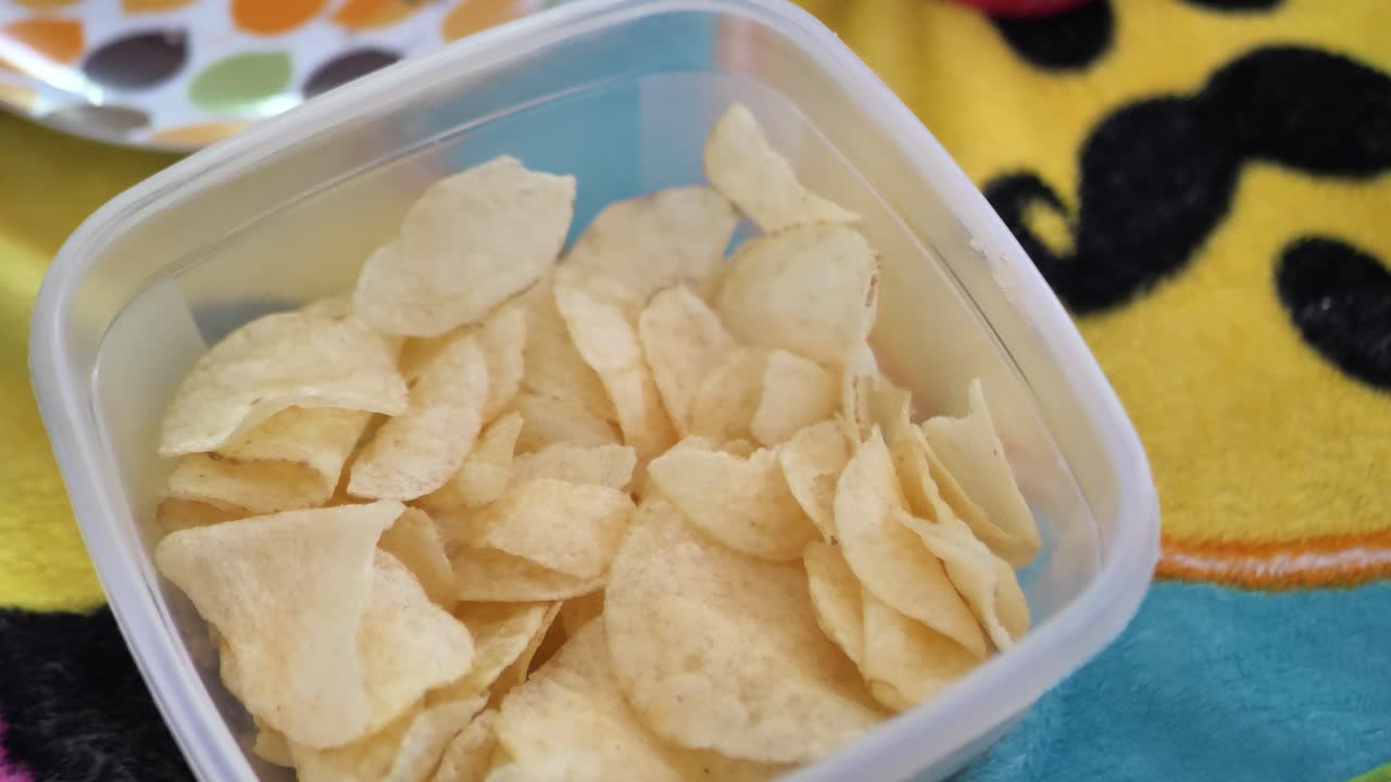 Due bambine caucasiche che mangiano patatine da una ciotola di plastica da asporto, con una coperta a tema emoji per terra sullo sfondo