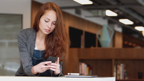 casually dressed young businesswoman checking mobile phone in modern open plan workplace