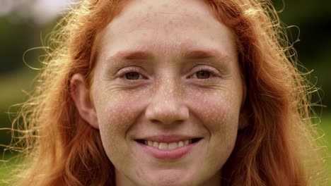 a young woman with red hair smiles warmly while standing outdoors in a lush green environment under the sun. her bright expression reflects joy and contentment.