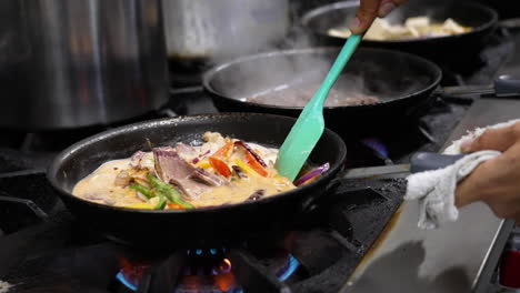 cinematic shot of crowded professional restaurant kitchen stove, line of sizzling skillets on range leading to cook stirring vegetables in sauce, slow motion close up slider 4k