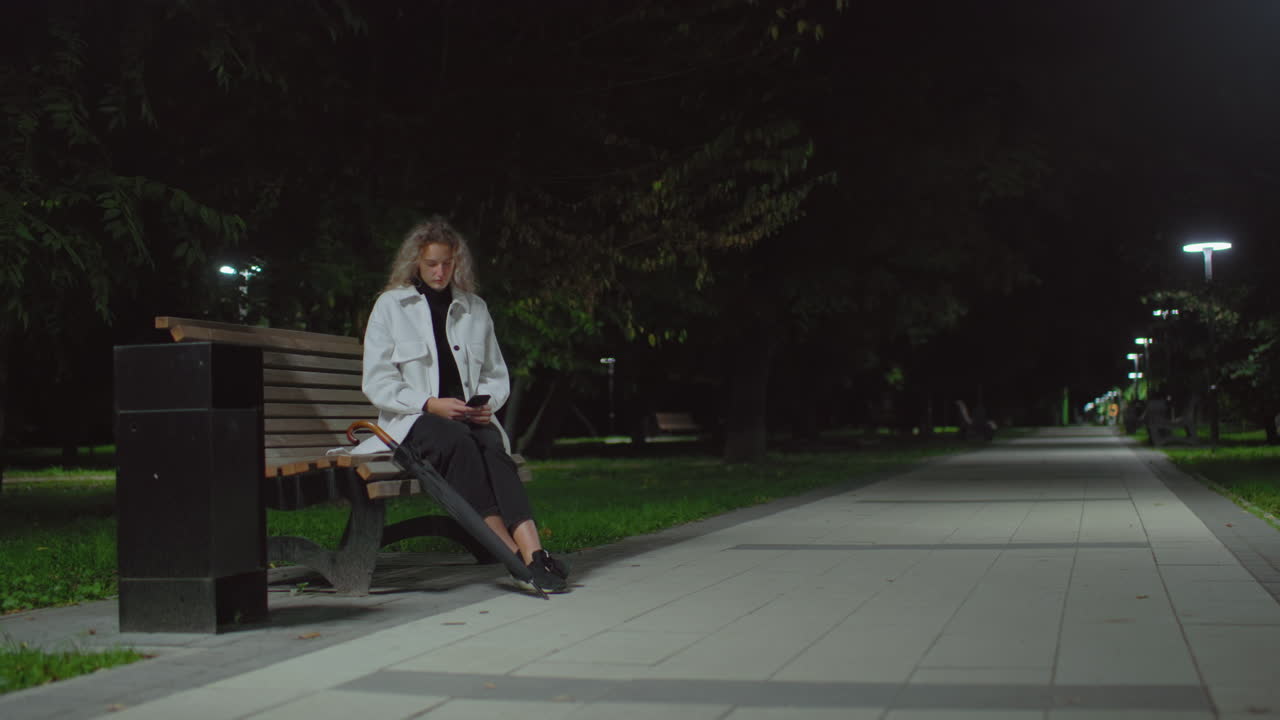 Young adult woman seated on wooden bench under streetlight at night using phone, surrounded by quiet park with trees, empty pathway, umbrella beside her, and calm ambient lighting in background