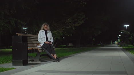 young adult woman seated on wooden bench under streetlight at night using phone, surrounded by quiet park with trees, empty pathway, umbrella beside her, and calm ambient lighting in background