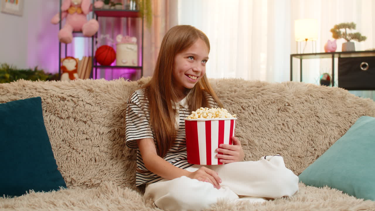 Child girl kid happily eats popcorn while watching cartoon on tv and laughing cheerfully on sofa