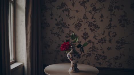 red rose resting in a white porcelain vase on a small round table, enhancing the elegance of a vintage style room with floral wallpaper and soft, inviting light