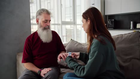 An-elderly-man-with-gray-hair-and-a-lush-beard-in-a-red-T-shirt-measures-his-blood-pressure-together-with-his-daughter-a-brunette-adult-girl-in-blue-pants-and-a-green-jacket-sitting-on-a-brown-sofa-in-a-modern-apartment