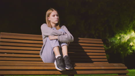 girl seated on wooden bench outdoors at night, gazing thoughtfully to the side, looking sleepy, resting her head gently on her shoulder under soft lighting with blurred background