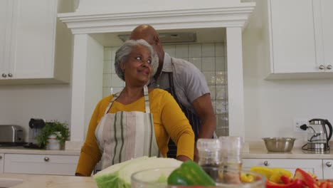 african american senior man tying apron from back to his wife in the kitchen at home