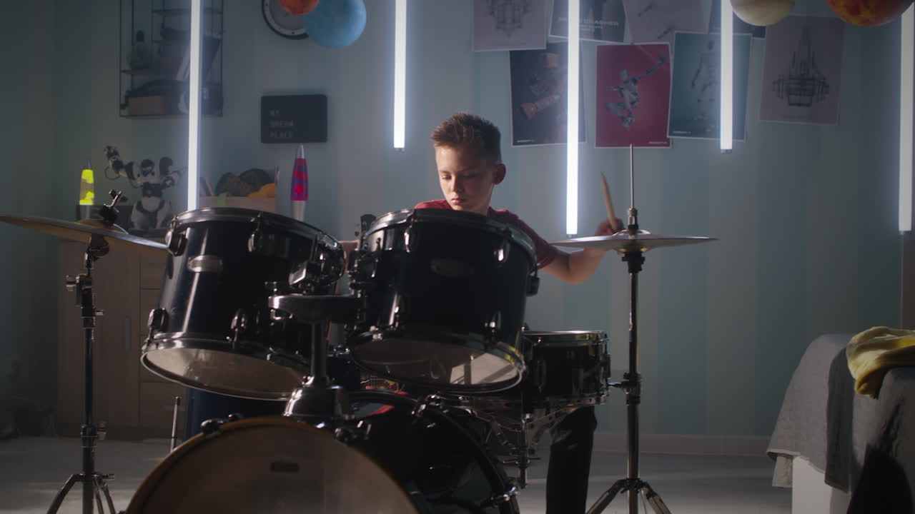 Teen Drummer Rehearsing in Bedroom Teenager in Casual Clothes Sitting Near Bed and Playing Drums during Rehearsal in Illuminated Bedroom at Home
