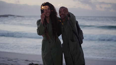 feliz pareja multiétnica en la playa bailando celebrando la víspera de año nuevo agitando bengalas disfrutando de una diversión tonta juntos en un tranquilo fondo marino junto al mar al atardecer