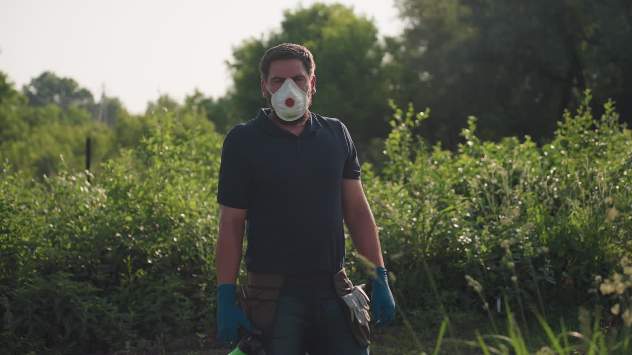 Un giardiniere con maschera protettiva e guanti blu si trova su un terreno agricolo lussureggiante, tenendo in mano una bottiglia spray verde, circondato da folto fogliame e alberi, e sembra fare una pausa durante il lavoro di fumigazione.