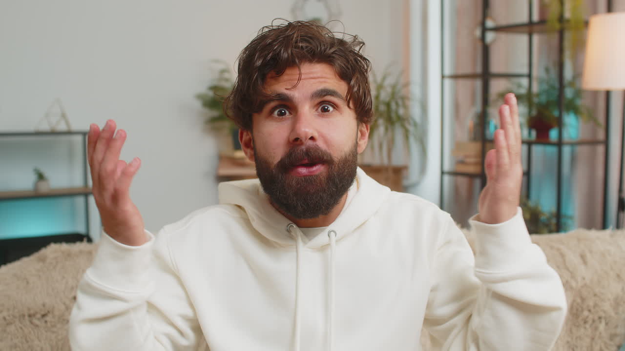 Wow emotion of bearded young indian man surprised looking at camera with big eyes sitting on sofa