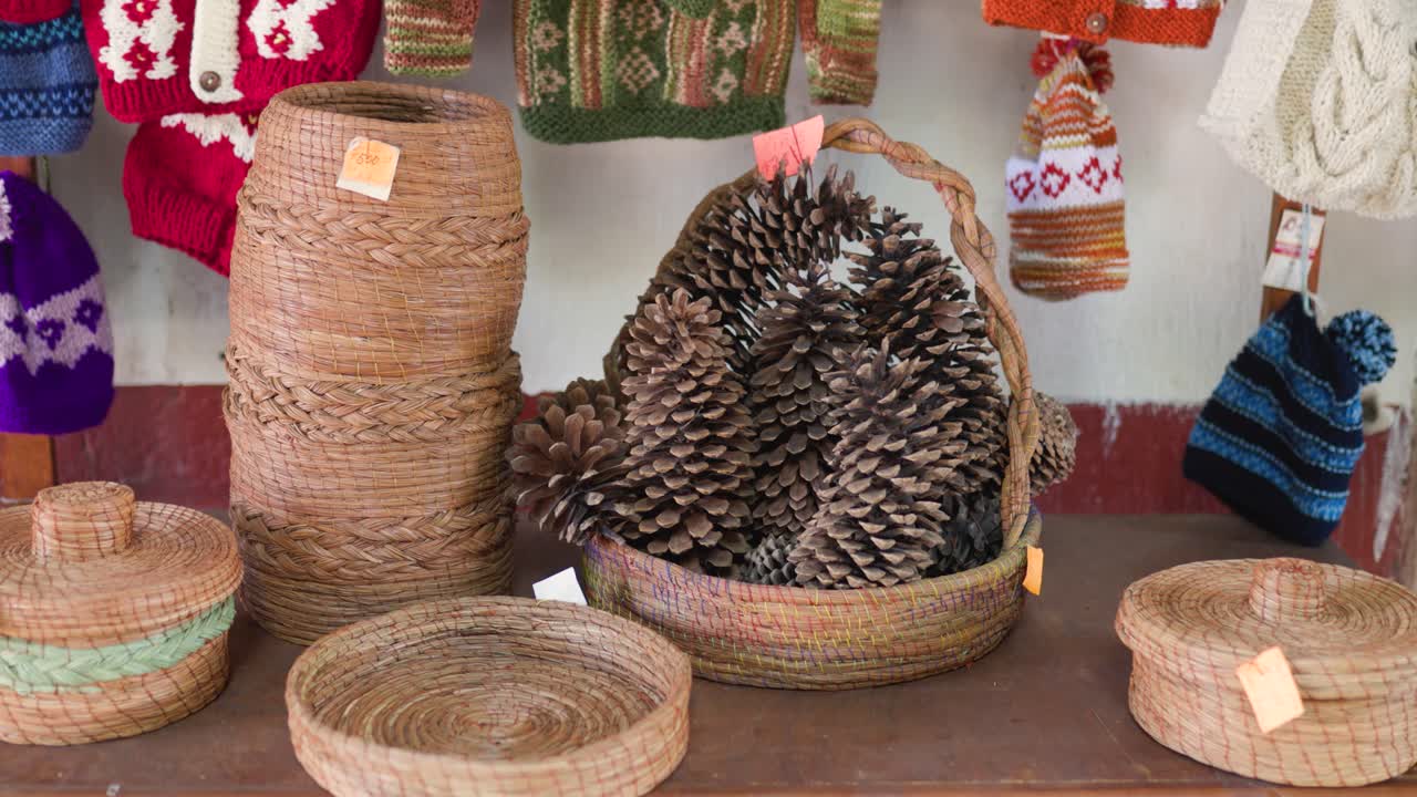 Large pine cones in a wicker basket on a wooden table surrounded by handwoven crafts and wool hats