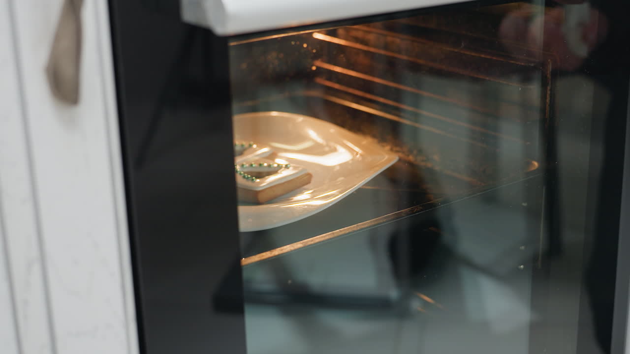 Close-up van twee versierde koekjes met witte glazuur en groene suikerparels op een wit bord in een oven, terwijl de ovendeur met doorzichtig glas wordt gesloten