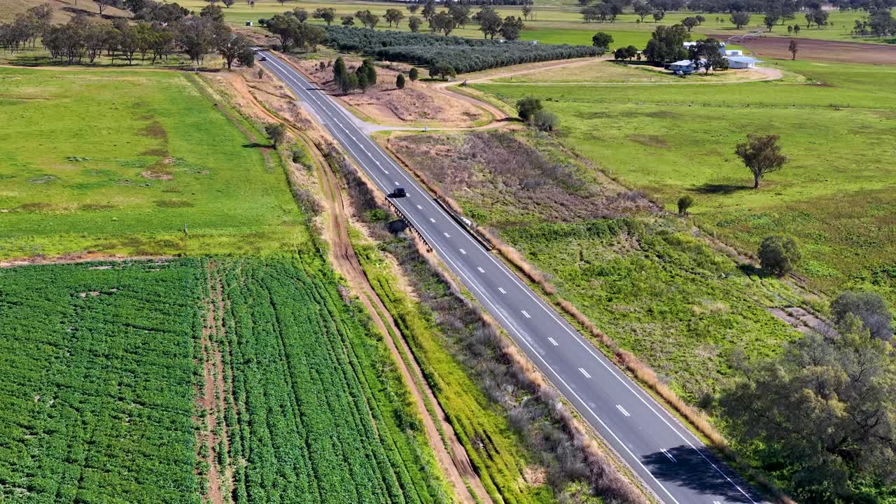 Drone footage captures a straight rural road cutting through green fields and rolling hills under bright daylight, with minimal traffic and stable overhead perspective
