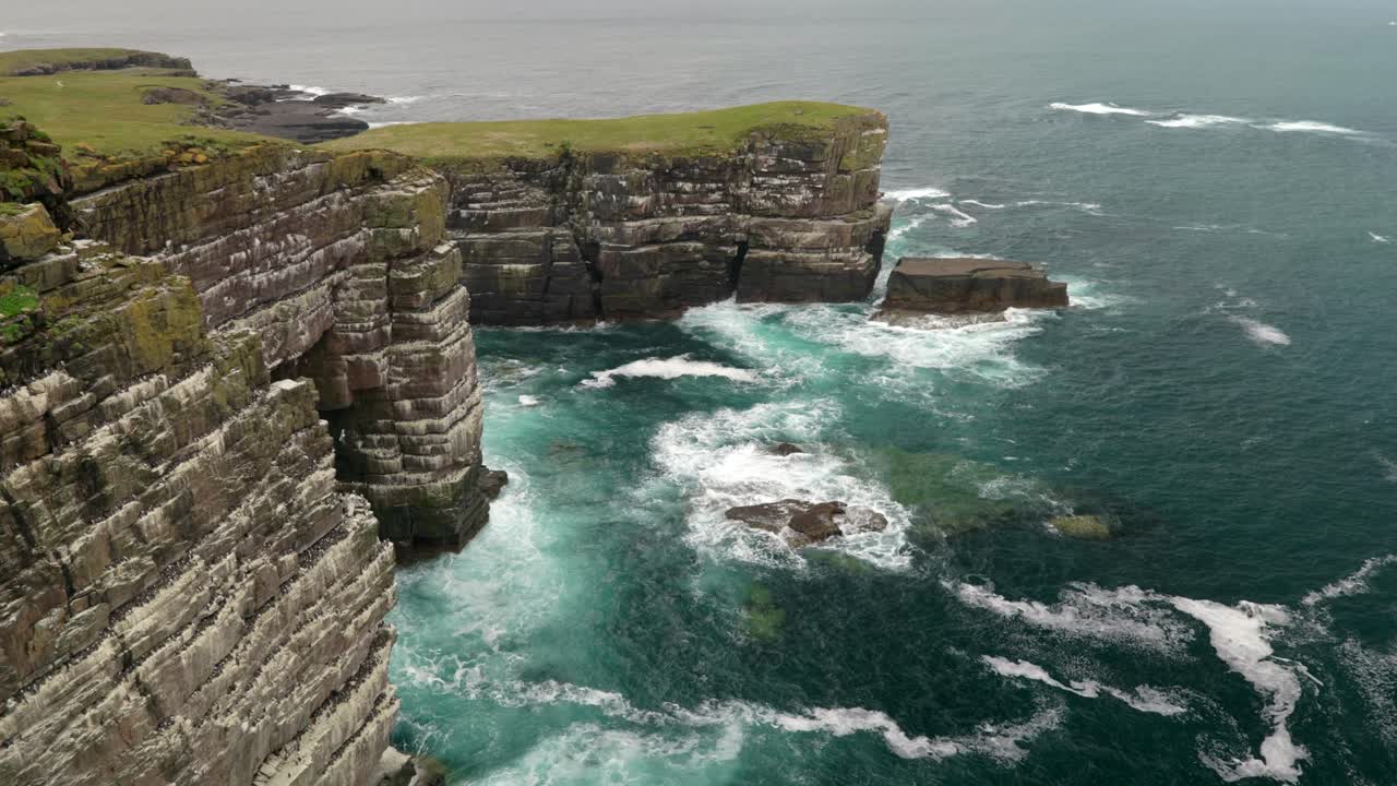Waves slowly crash in the deep teal coloured ocean over rocks and against an steep sea cliff covered in seabirds while seabirds fly around the cliffs of a colony of guillemots