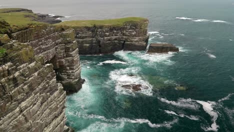 waves slowly crash in the deep teal coloured ocean over rocks and against an steep sea cliff covered in seabirds while seabirds fly around the cliffs of a colony of guillemots