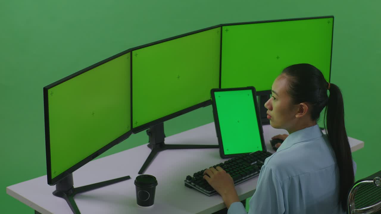 vista lateral de una mujer asiática trabajando con una tableta y varios monitores de computadora con una maqueta de pantalla verde en un estudio de pantalla verde