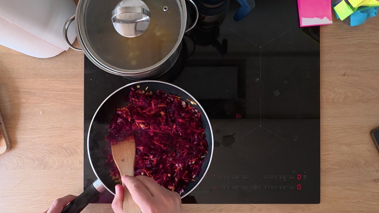Overhead close-up shot of a person stirring vibrant vegetables in a black pan on an electric stove. The modern kitchen setting showcases a wooden spatula and organized countertop.