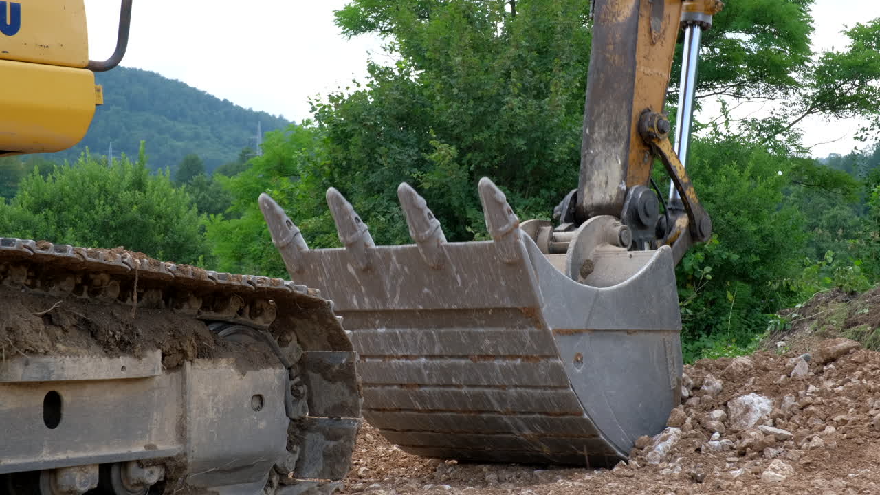 A close view shows a construction vehicle with a track and a large shovel Dirt and rocks surround the machine Trees are in the background