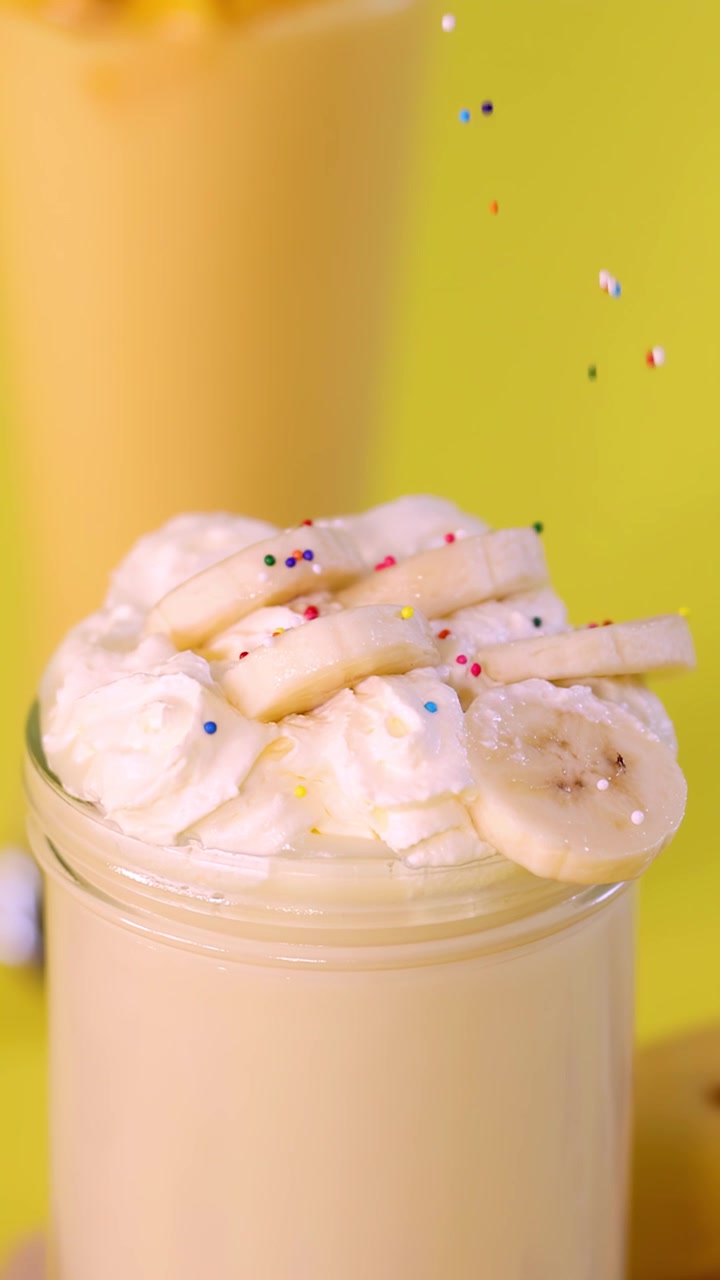 Frullato di banana guarnito con panna montata e fette di banana, con codette colorate che cadono, sotto una luce intensa.