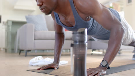 focused african american man doing push ups next to water bottle in sunny living room, slow motion