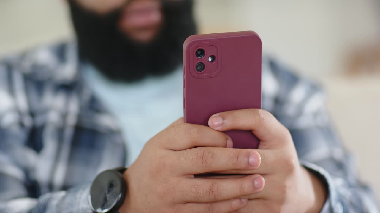 Uomo adulto sul divano che avvicina un telefono bordeaux, tenendolo con due mani, scorre il feed, orologio visibile.