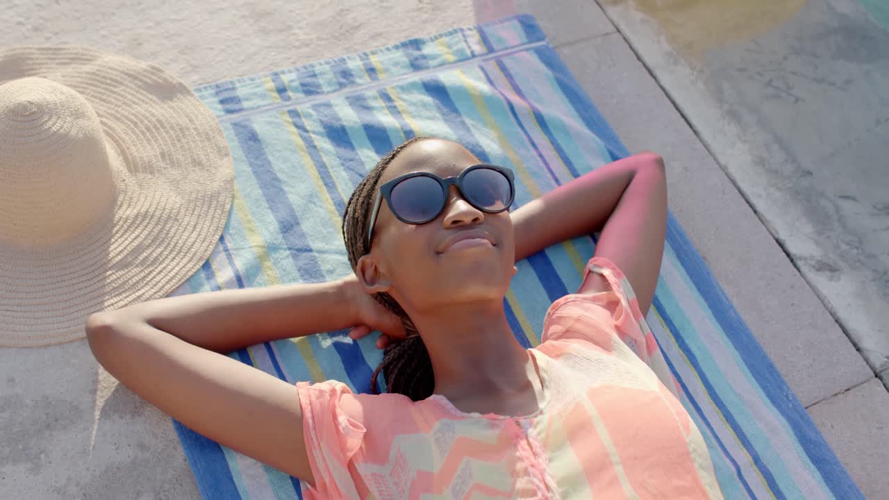Relaxed, smiling african american woman in sunglasses sunbathing on towel by pool, in slow motion
