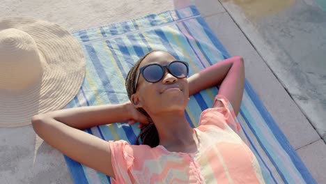 Mujer-Afroamericana-Relajada-Y-Sonriente-Con-Gafas-De-Sol-Tomando-El-Sol-En-Una-Toalla-Junto-A-La-Piscina,-En-Cámara-Lenta