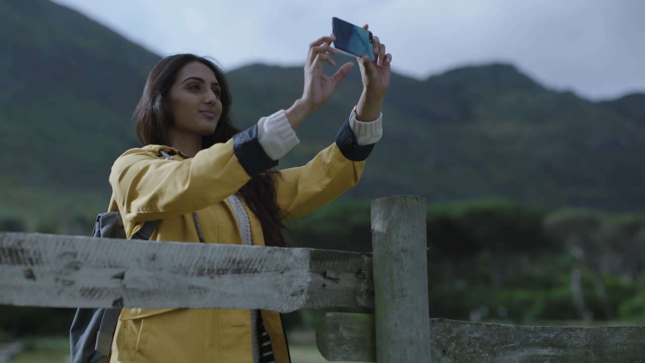 beautiful young indian woman taking photo using smartphone camera technology enjoying sharing adventure travel hike photographing green countryside outdoors