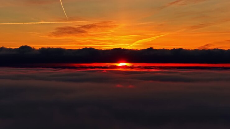 Alba arancione infuocata che squarcia lo strato di nubi scure sopra un orizzonte soffice e nebbioso visto da alta quota