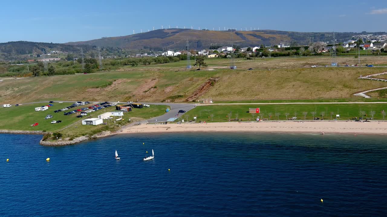 gente en la playa y navegando en el lago con el pueblo detrás y las turbinas eólicas en el fondo de las montañas