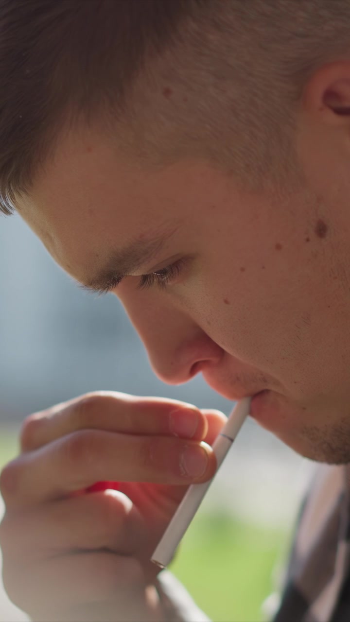 Primo piano di un giovane uomo che tiene e usa una sigaretta elettronica all'aperto con luce naturale soffusa. La mano del soggetto è visibile mentre afferra il dispositivo, mentre lo sfondo mostra edifici urbani sfocati