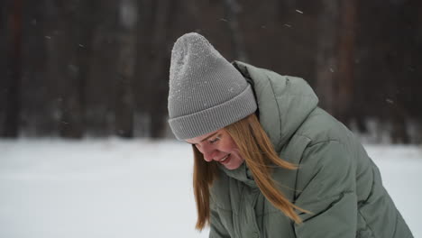 young woman with long blonde hair wearing gray beanie and green puffer jacket looks downward while smiling subtly in snowy forest setting, snowflakes resting on hat and falling around her in cold serene