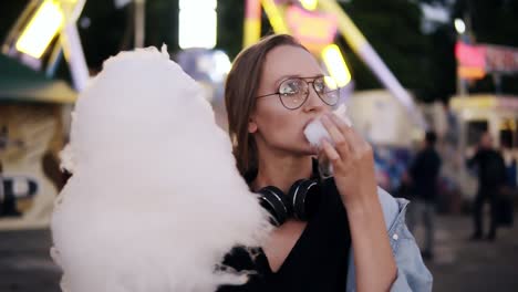 and attractive girl walking in the amusement park, eats white cotton candy. girl wearing casual clothes, headphones on her neck