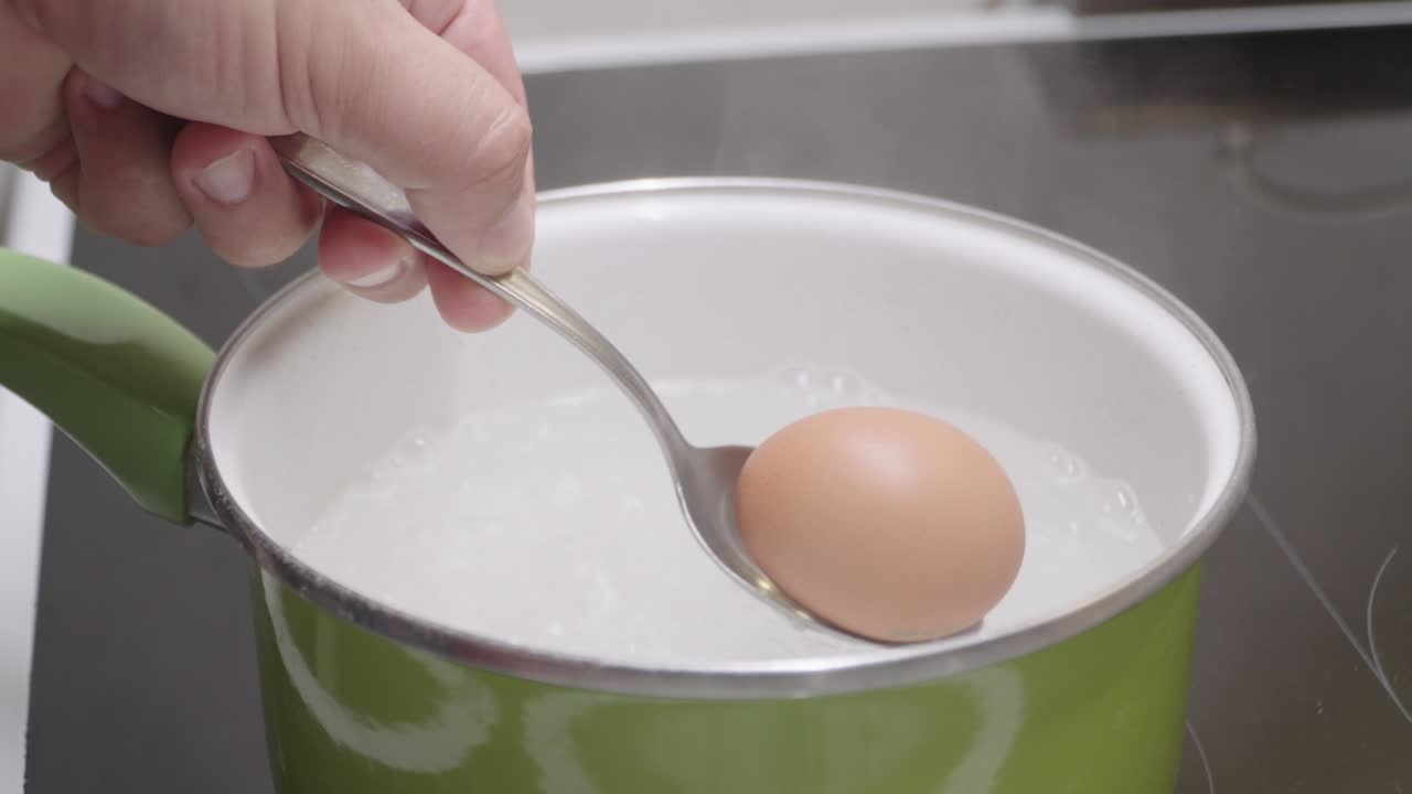 Egg lowered into boiling water with spoon in saucepan close up cooking process