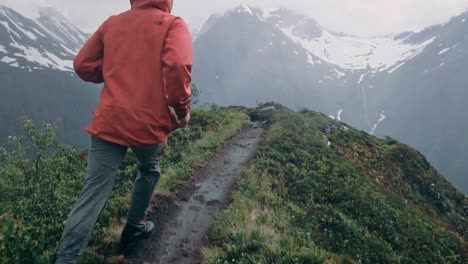 slow motion shot of man in a red jacket running over the peak of a moutain ridge, dashing through bushes surroudned by snow white mountains.