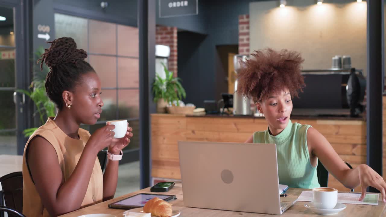 Femmes afro-américaines en pleine planification dans un café, l'une tapant sur une tablette, échangeant des documents et examinant un ordinateur portable.