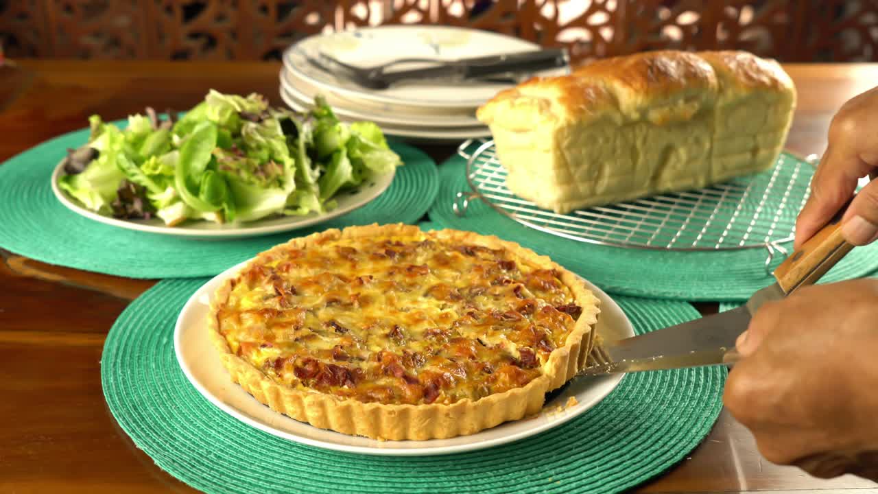 Senior hand using metal cake serving shovel and knife taking a piece of homemade green pea and bacon quiche from white ceramic dish.