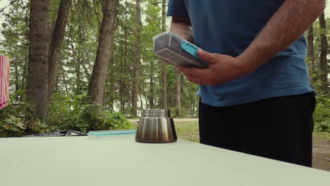 man spoons coffee grounds into base of moka pot on table at campsite