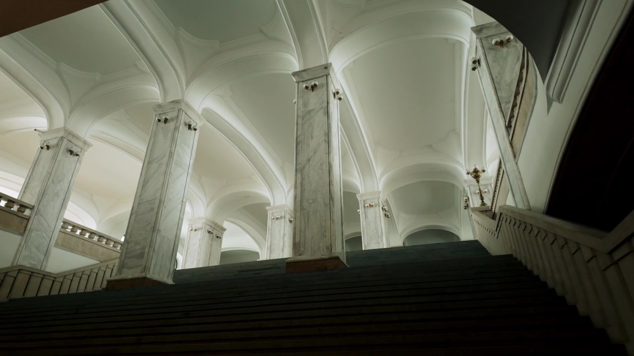 Ascending staircase within Holy Trinity Cathedral in Tbilisi, Georgia. Features elegant marble pillars, graceful archways, and dim lighting creating a moody ambiance.