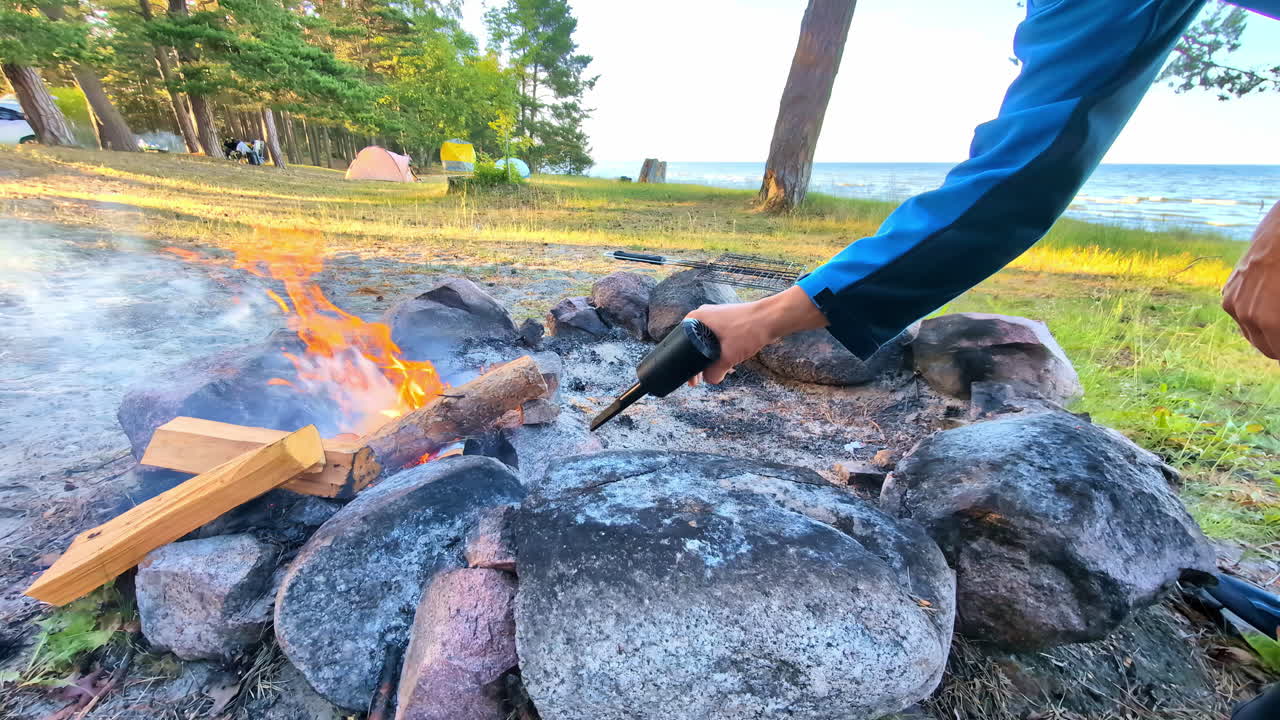 A person blows air on a camping fire by the lake, making the flames grow bigger and brighter.