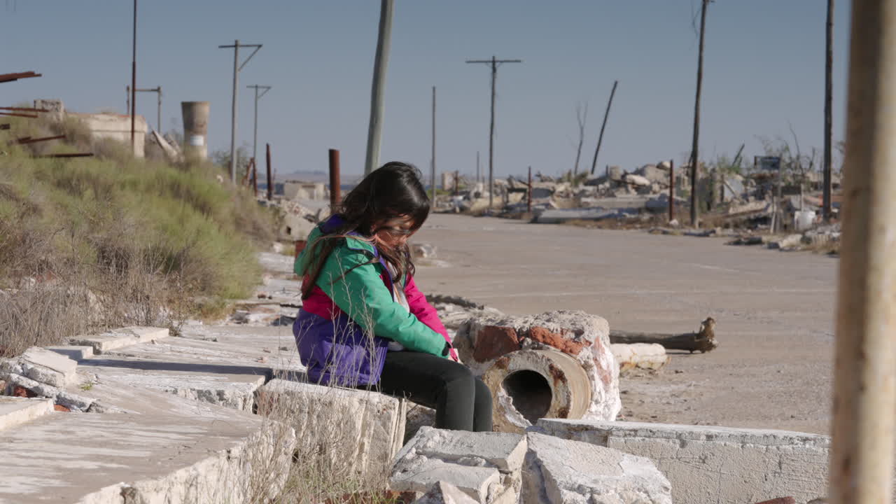 Young girl sitting alone on a sidewalk in a destroyed city, surrounded by ruins, symbolizing war aftermath, loss, vulnerability, and civilian impact