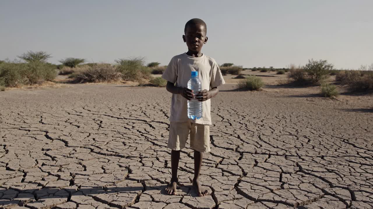 niño africano esperanzado de pie en tierra seca y agrietada, agarrando una botella de plástico de agua limpia, simbolizando la lucha por la supervivencia en medio de la sequía y el cambio climático