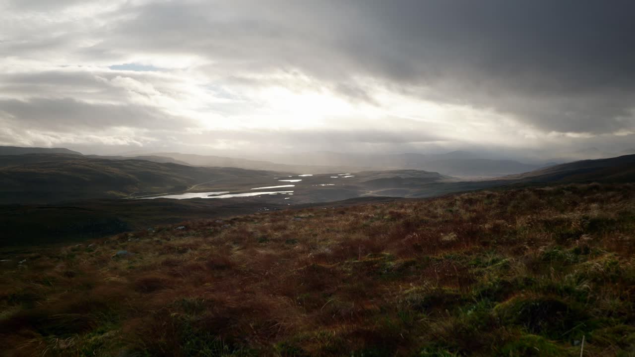 los vientos rápidos soplan hierba en primer plano contra un paisaje oscuro de montañas y lagos en las tierras altas del noroeste de escocia mientras rayos de luz perforan el cielo