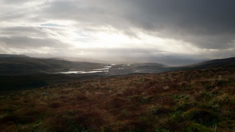fast winds blow grass in the foreground against a dark landscape of mountains and lochs in the north west highlands of scotland as shafts of light pierce the sky
