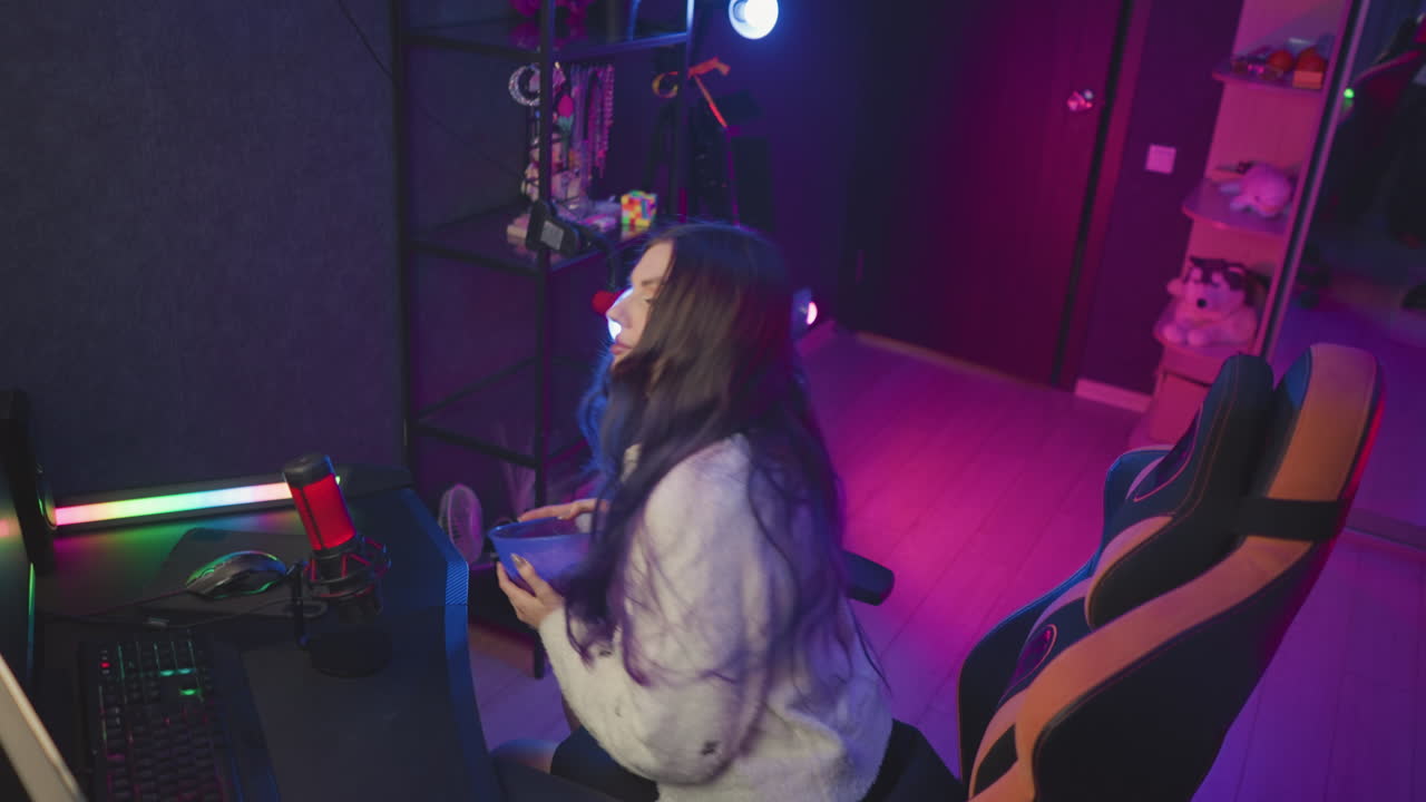 Young woman holding bowl of popcorn walks toward ergonomic chair in vibrant gaming studio filled with colorful lighting preparing to sit and adjust seating for comfortable viewing experience