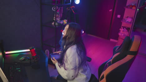 young woman holding bowl of popcorn walks toward ergonomic chair in vibrant gaming studio filled with colorful lighting preparing to sit and adjust seating for comfortable viewing experience