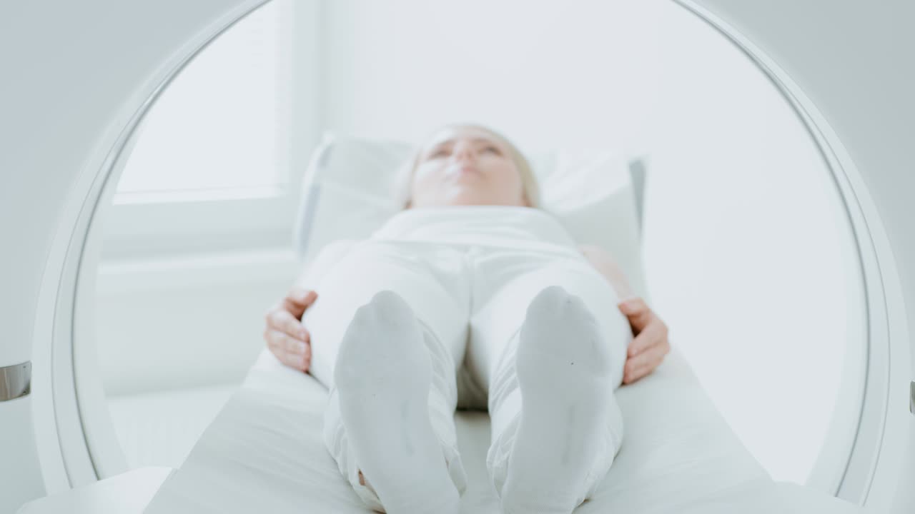 Primo piano di una paziente sdraiata su una macchina per TAC o risonanza magnetica, il lettino si muove all'interno della macchina mentre scansiona il suo corpo e il suo cervello. In un laboratorio medico con attrezzature ad alta tecnologia. Si allontana dalla telecamera.