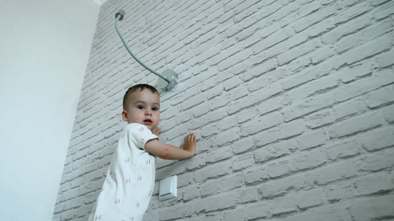 Adorable Caucasian toddler reaching to the lamp on the wall. Baby boy plays with light at home. Low angle view.
